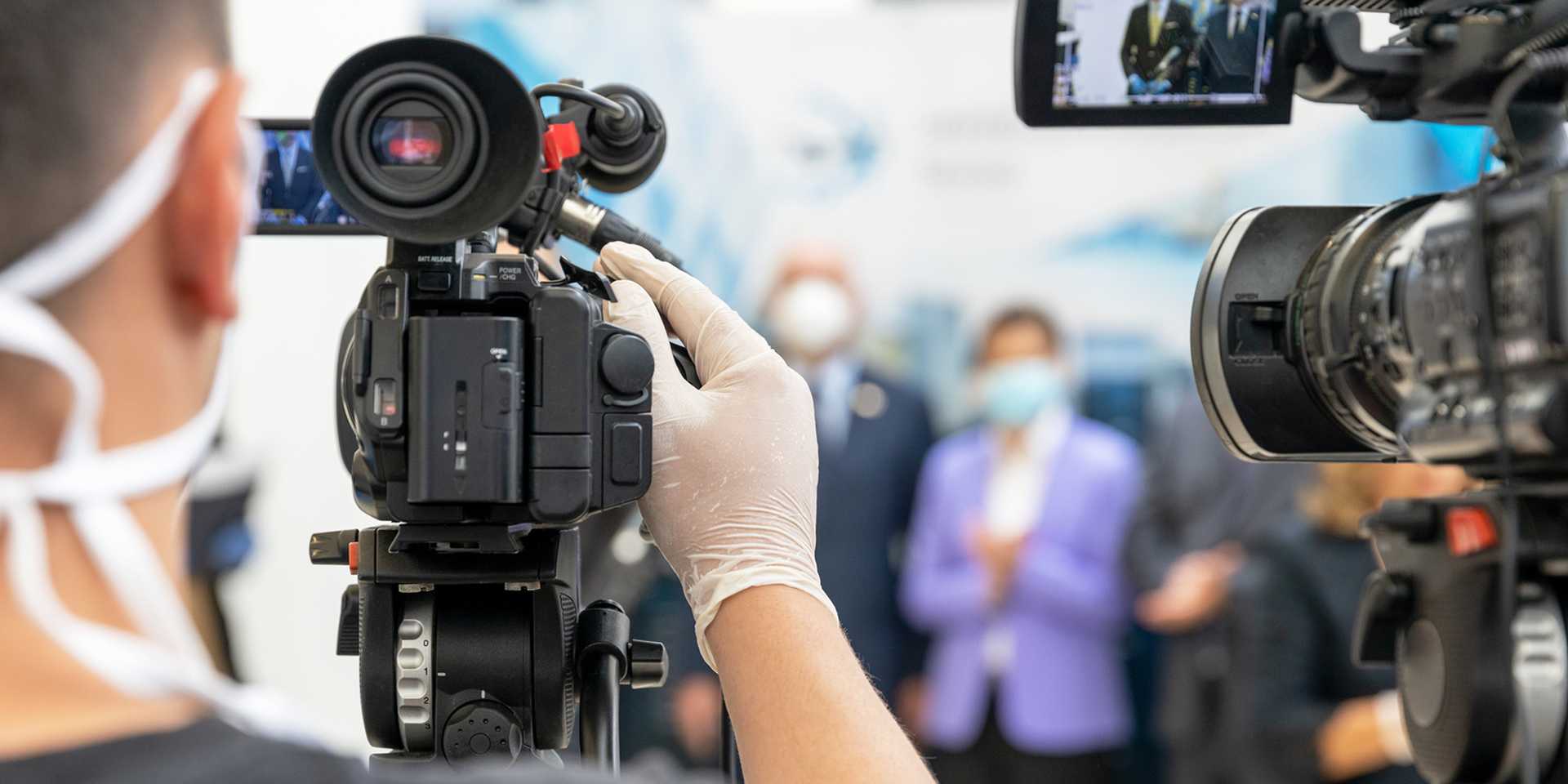 A cameraman in mask and gloves filming a press conference where people are wearing face masks