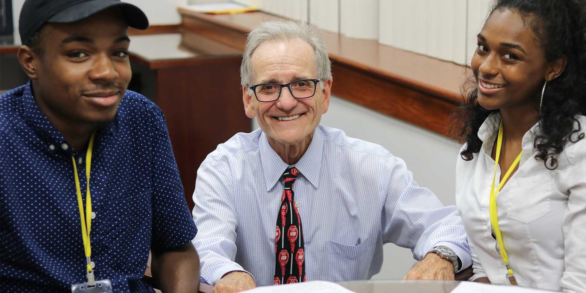 Two teens sit at a table while an older man kneels between them smiling at the camera