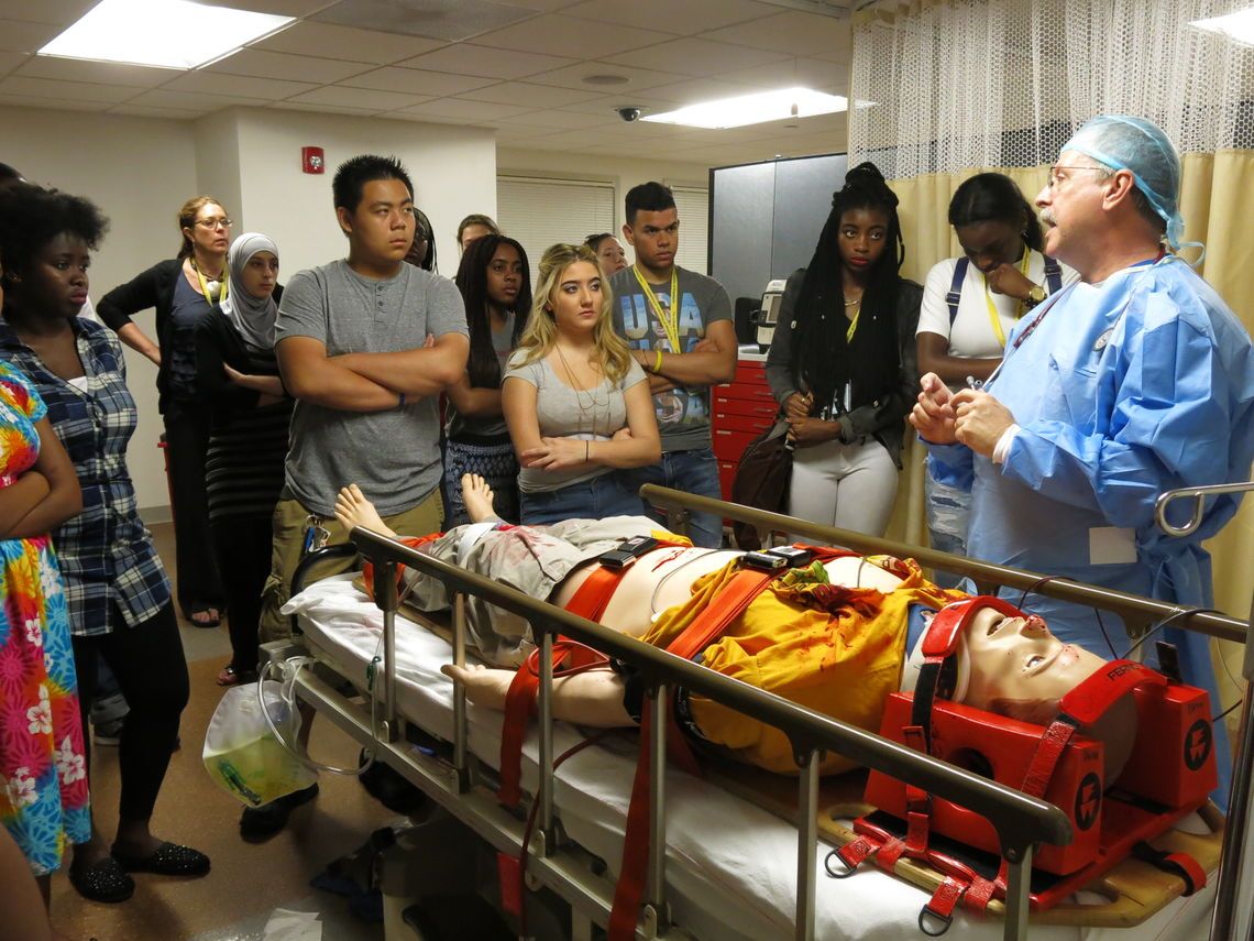 Students stand around a gurney holding a medical dummy. A doctor in scrubs addresses the students.