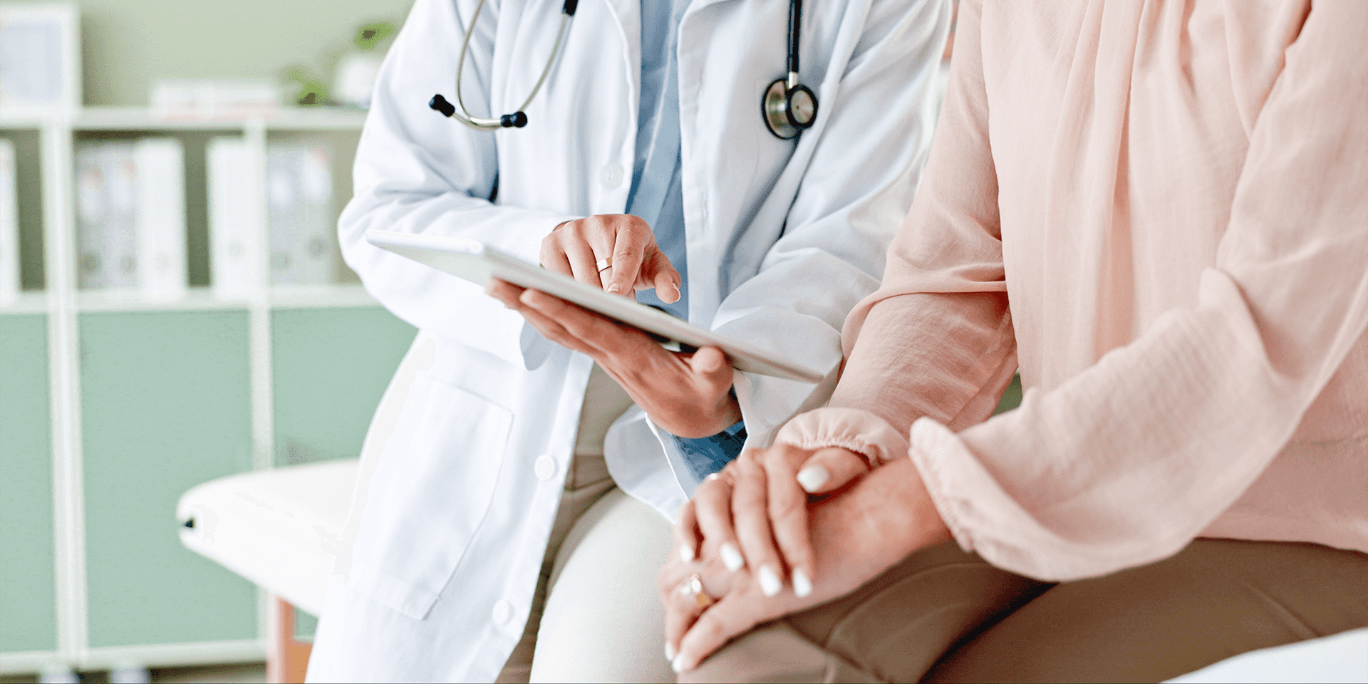 A doctor in a white coat sits next to a woman in a pink, long sleeved shirt. The doctor holds a medical chart and points to it