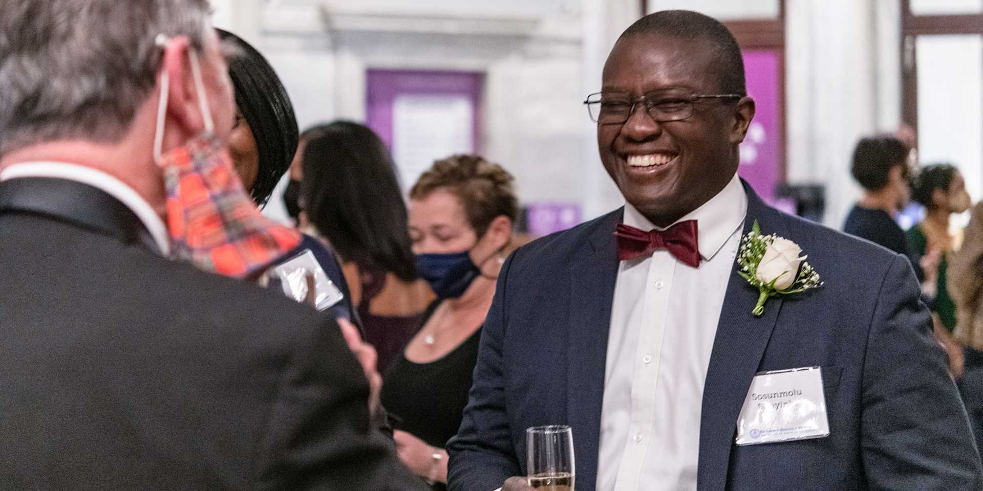 Man in suit with corsage smiling and drinking champagne while speaking to another man