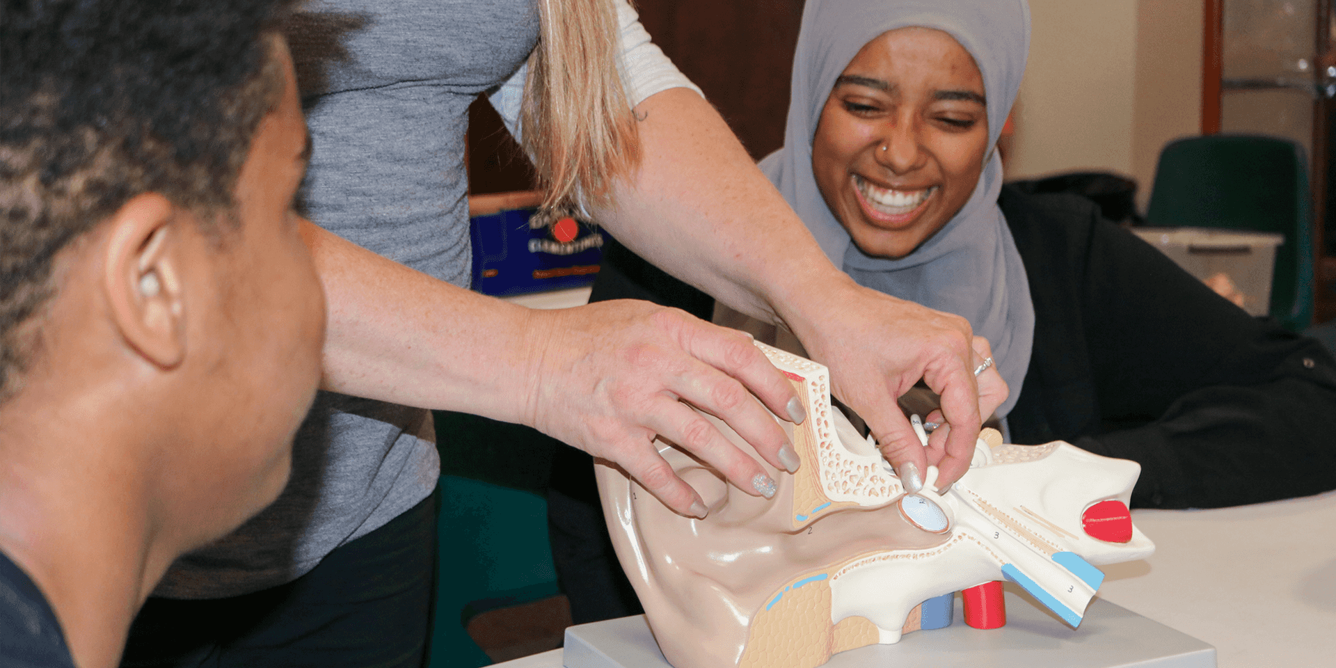 Two students seated at a table while an instructor displays an anatomical model of the human ear