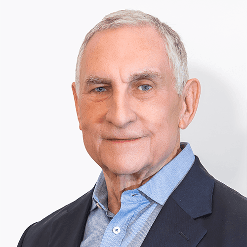 Headshot of John Hansen-Flaschen, a man with short gray-white hair and blue eyes. He wears a light blue button-down shirt under a navy blazer, and stands against a plain white wall.