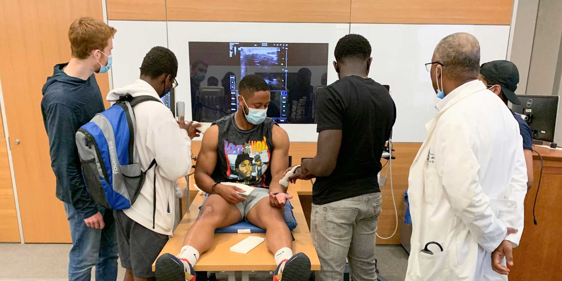 A young Black man sits on a table while three other young men demonstrate medical devices, while a doctor oversees.