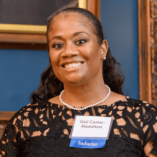 Photograph of a Black woman with dark hair pulled away from her face. She wears a black lace dress and a pearl necklace. A nametag pinned to her dress reads "Gail Carter-Hamilton" and a blue ribbon says "Inductee"