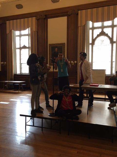 Students in the KArabots Junior Fellows Program stand on a stage in Mitchell Hall at the College of physicians of Philadlephia. They receive small trophies for competing in an educational game show.